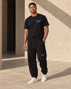 Model wearing the black Signature Tee with matching black sweatpants in a stone courtyard.